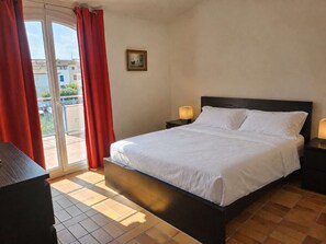 Una habitación de hotel con una cama, dos mesitas de noche con lámparas, una puerta de vidrio corredera con cortinas rojas y un cuadro pequeño en la pared.