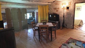 A rustic dining area with a wooden table and chairs, a television on a stand, and a stone wall. - Homerez - Flat in Feliceto (Feliceto)