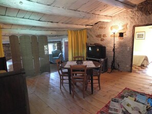 Un coin repas rustique avec une table et des chaises en bois, un téléviseur posé sur un meuble et un mur en pierre.