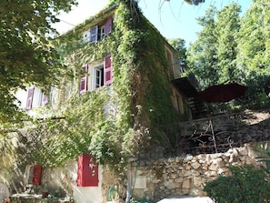 Un bâtiment rustique en pierre, aux murs recouverts de lierre et aux volets rouges, doté d’un escalier en pierre et d’une rampe en métal.