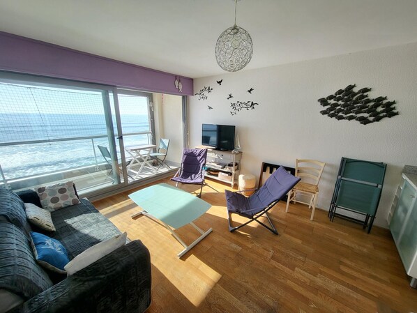 A living room with a sofa, a small table, a TV, and a balcony with a view of the sea. - Studio with balcony, WIFI, and parking near the beach (Les Sables-d'Olonne)