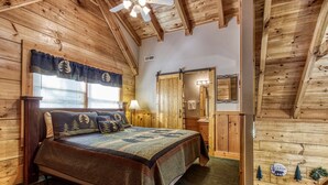 Ein gemütliches Schlafzimmer im Blockhausstil mit einem großen Bett, Holzwänden, einem Fenster mit Aussicht und einem Deckenventilator.