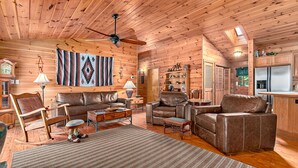 A cozy log cabin interior with a living room, leather sofas, a coffee table, and a dining area. - Secluded, Close to Town, Hot Tub, Fire Pit, WiFi, Pets Ok! (Fairview)