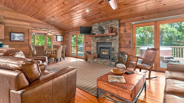 A spacious living room with leather furniture, a stone fireplace, and large windows overlooking a deck. - Secluded, Close to Town, Hot Tub, Fire Pit, WiFi, Pets Ok! (Fairview)