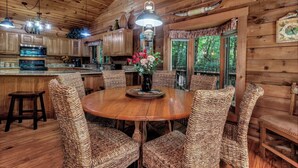 A rustic wooden cabin interior with a round dining table surrounded by wicker chairs, a kitchen area with wooden cabinets, and a hanging light fixture. - Secluded, Close to Town, Hot Tub, Fire Pit, WiFi, Pets Ok! (Fairview)