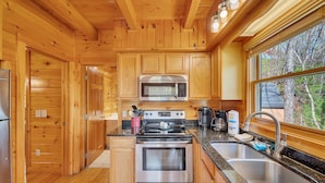 Cocina de cabaña de madera con refrigerador de acero inoxidable, microondas y horno, encimera de granito y ventana con vista a árboles.