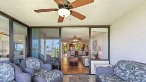 A spacious living area with a ceiling fan, sliding glass doors leading to a kitchen and dining area, and a large window providing natural light. - Ocean view, Country Club Villas 224, No stairs, 2nd floor unit. (Kailua-Kona)