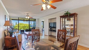 A spacious living room with a glass dining table, wooden chairs, a ceiling fan, and a large window offering a view of the outdoors. - Ocean view, Country Club Villas 224, No stairs, 2nd floor unit. (Kailua-Kona)
