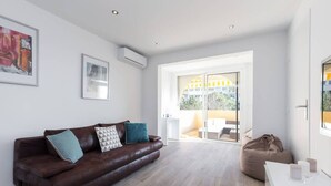 A modern living room with a brown leather sofa, a white coffee table, and a beige bean bag. There are two framed artworks on the wall and a large window providing a view of the outside. - Haven of Peace: 1-Br Terrace with Sea View & Pool (Cannes)