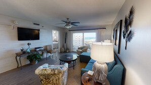 A modern living room with a flat-screen TV, a ceiling fan, a sofa, a coffee table, and a view of the ocean. - Sunset Views & Summer Beach Vibes – Islander 713 (Destin)