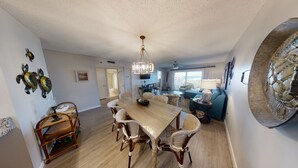 A spacious living area with a wooden dining table, wicker chairs, a blue sofa, and a large decorative mirror. - Sunset Views & Summer Beach Vibes – Islander 713 (Destin)