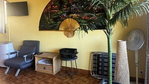 A room with a sofa, a small table, a fan, a decorative wall hanging, and a large plant.