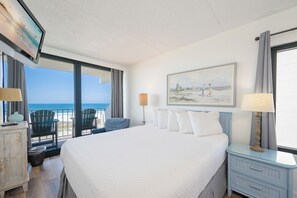 A hotel room with a large bed, a television, a painting, and a view of the beach.