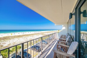 A balcony with a view of the beach, outdoor seating, and a clear sky.