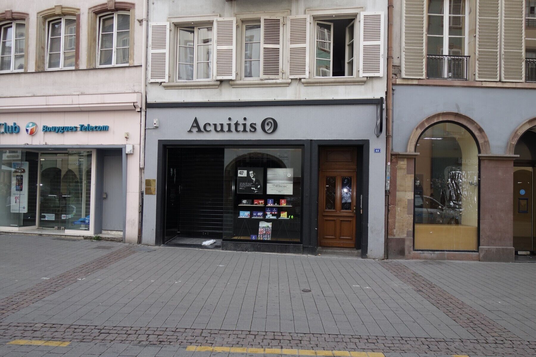 Een winkelgevel met een bord waarop 'Acuitis' staat en een gesloten luik.