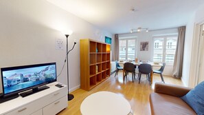 Un salon moderne doté d’un téléviseur à écran plat, d’étagères en bois, d’un coin repas avec des chaises et d’une grande fenêtre agrémentée de rideaux.