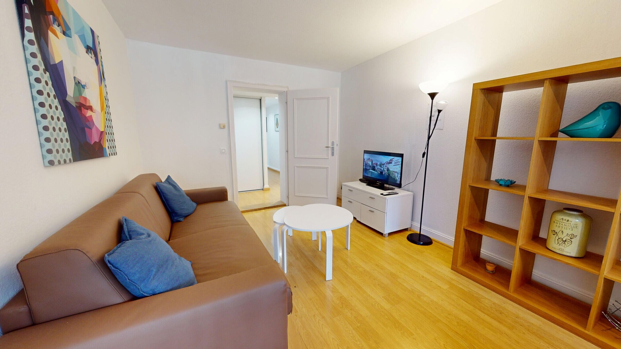 Ett vardagsrum med en brun soffa, ett vitt soffbord, en tv på ett vitt tv-bord och en bokhylla i trä.