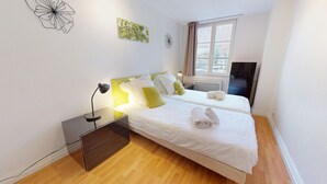 A hotel room with two beds, a desk with a lamp, a window with curtains, and artwork on the walls.