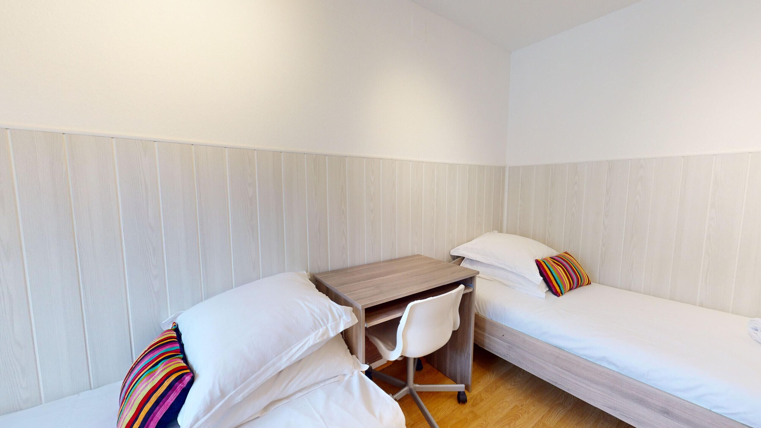 A small, single-room accommodation with a wooden desk, a chair, and two beds.