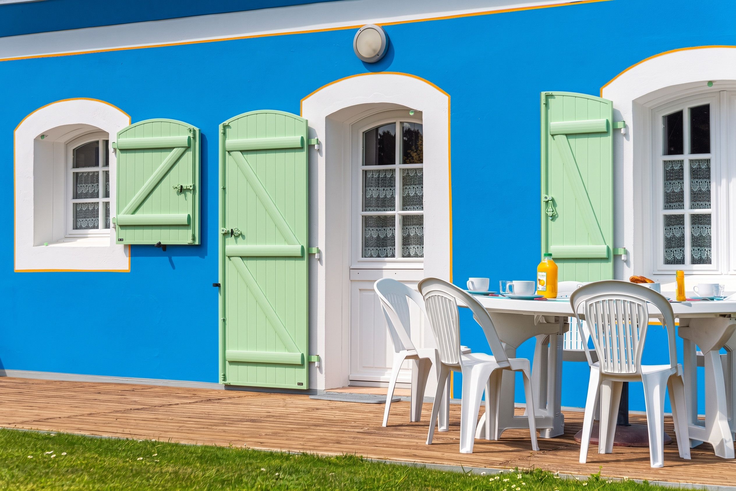 A building with a blue exterior, white trim, and green shutters. There is a table with chairs and a bottle of orange juice.
