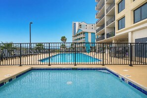 A swimming pool with a black metal fence, surrounded by a building with balconies and a clear blue sky.
