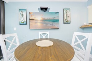 A dining area with a round wooden table, white chairs, and artwork depicting sailboats and a beach scene. - Island Winds 1 BR *Gulf Front* (Gulf Shores)