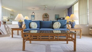 A living room with a blue sofa, two side tables with lamps, a glass coffee table, a ceiling fan, and a view of the kitchen through a large mirror.