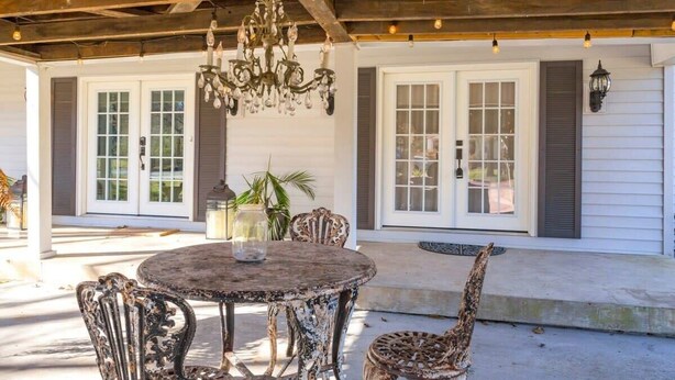 Une terrasse extérieure avec une table ronde et des chaises, un lustre et une maison aux murs recouverts de bardage blanc et aux volets fermés.