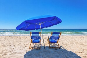 Deux chaises de plage sous un parasol bleu, sur une plage de sable, avec l’océan en arrière-plan.