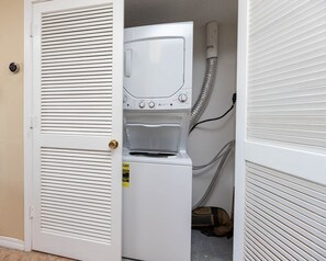 A laundry room with a front-loading washer and dryer, a door with a brass handle, and a visible hose. - Royal Palms 1BR 2BA! Condo 1207 ~ (Gulf Shores)