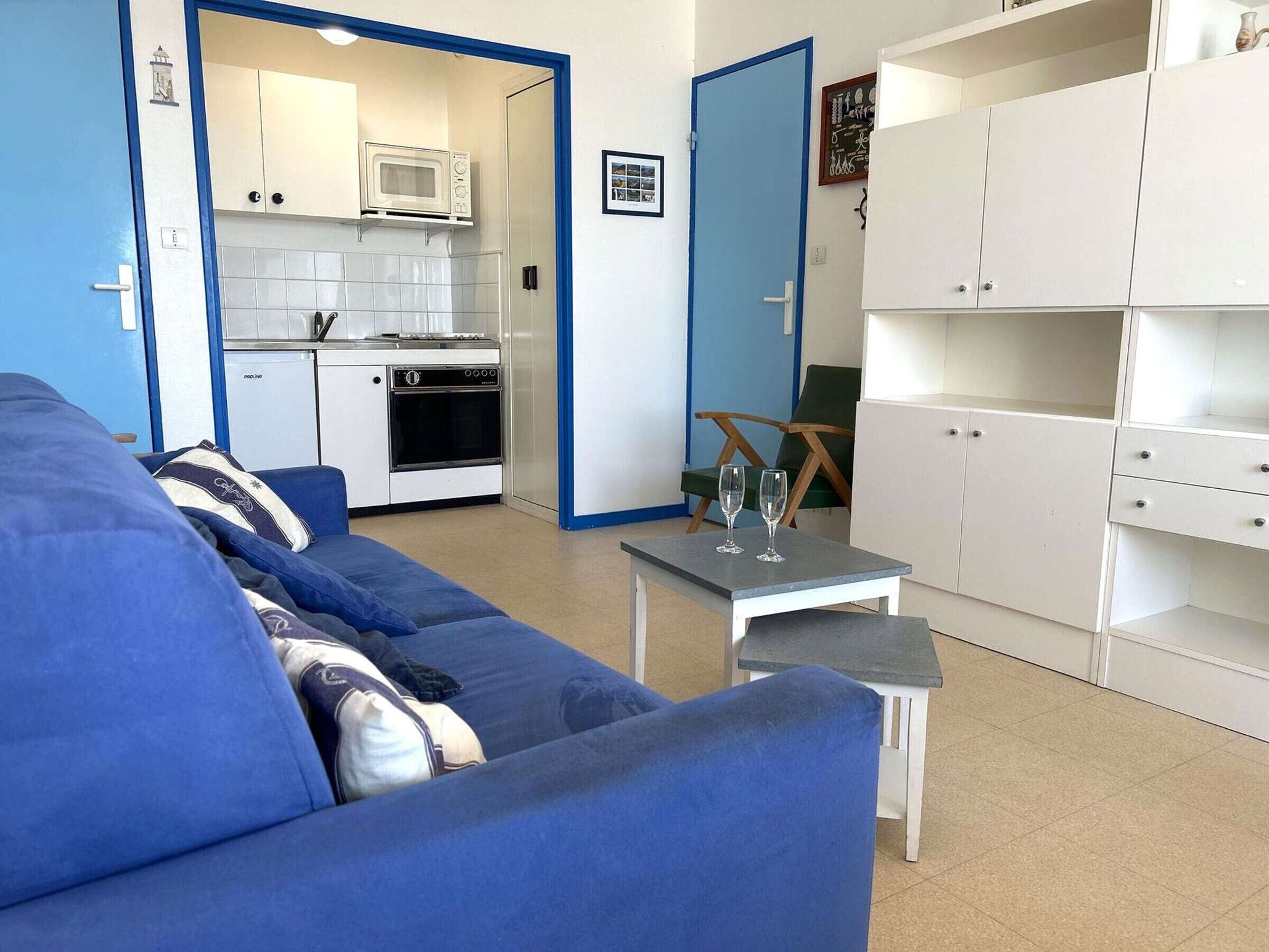 Ein gemütlicher Wohnbereich mit Küchenzeile, einem blauen Sofa, einem kleinen Tisch mit Gläsern und weißen Schränken.