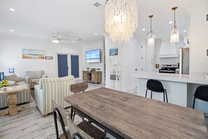 A modern living room with striped sofas, a wooden dining table, and a kitchen area with white cabinets and a stainless steel refrigerator.