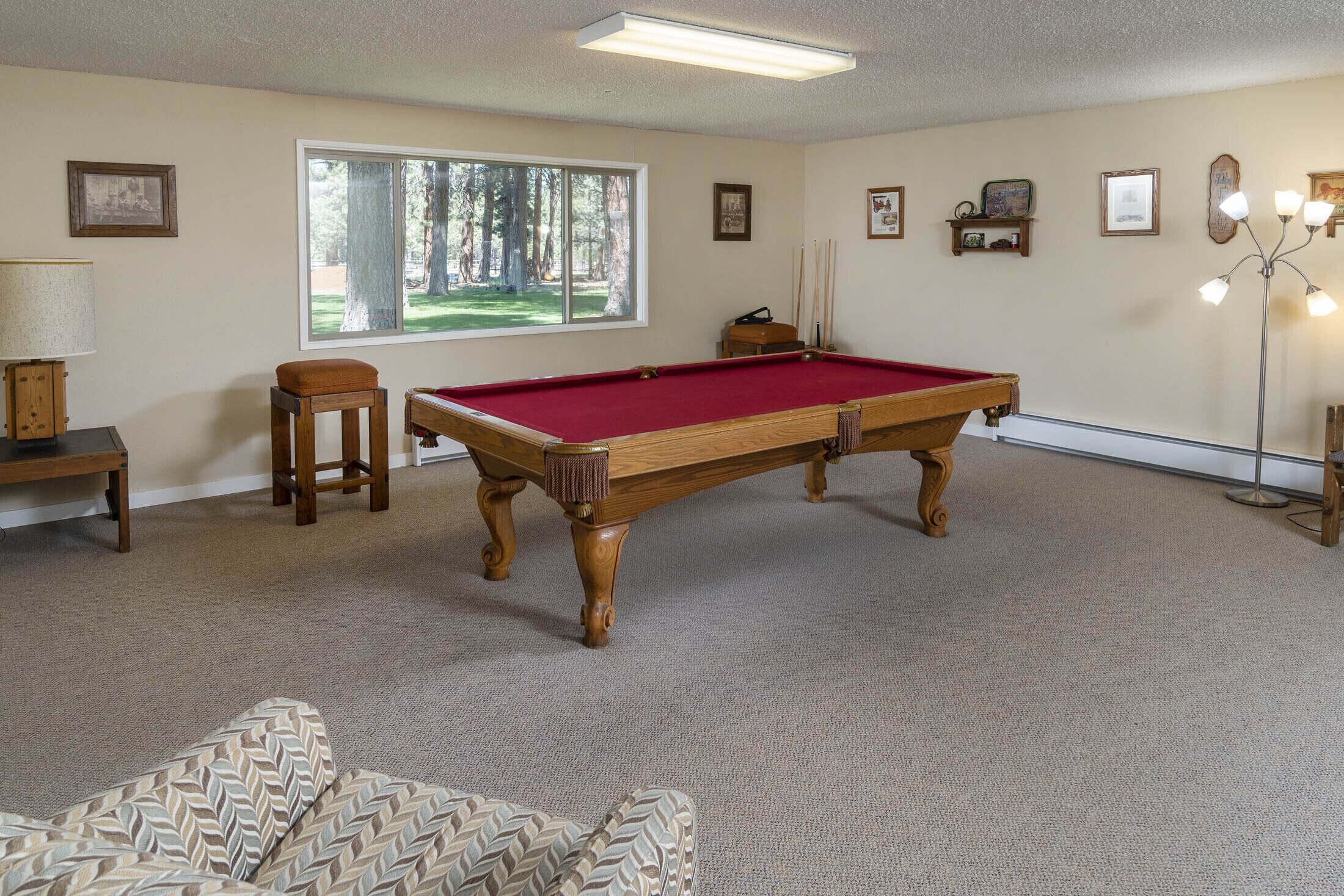 A room with a pool table, a sofa, a window with a view of trees, and a lamp.