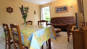 A dining area with a table covered in a floral tablecloth, wooden chairs, a vase with flowers, a ship's wheel decoration, a framed picture on the wall, and a brown sofa. - Holiday home 4 pers, private garden, in La Hague (Vasteville)