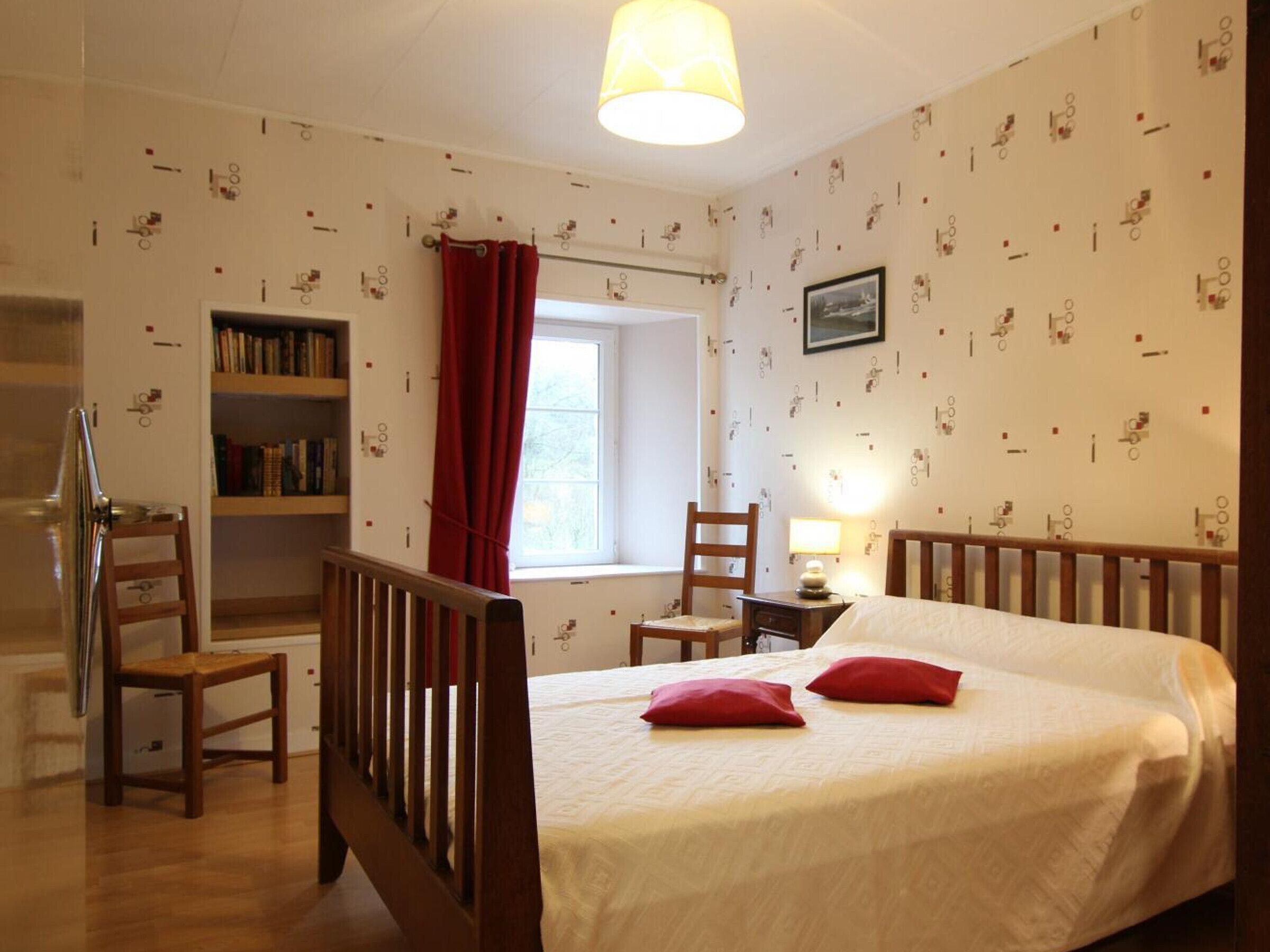 Een slaapkamer met een bed, een boekenplank, een stoel, een bureau met een lamp en een raam met rode gordijnen.