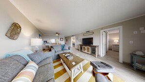 A modern living room with a sofa, coffee table, and a TV. There is a kitchen in the background and a door leading to another room. - 🌅 Islander #301 – Luxury Beach Living, Steps to the Sand! 🌊🐬 (Destin)