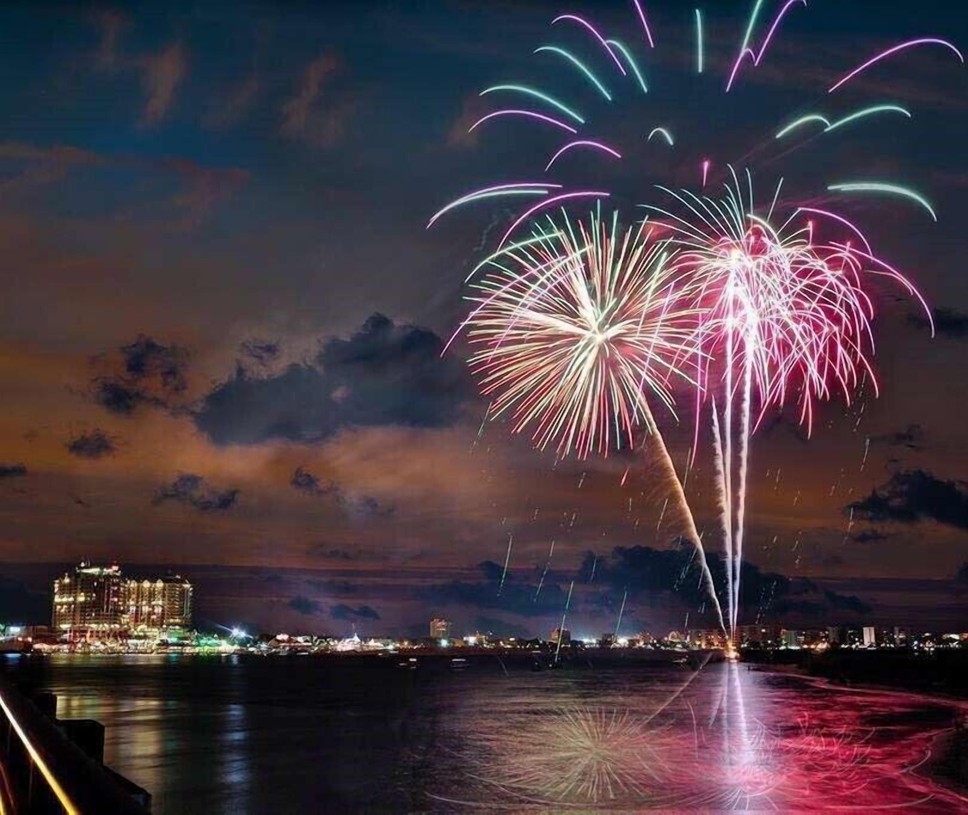 Een nachtscène met vuurwerk boven het water, een stadsachtergrond en een hotel met verlichting.