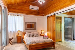 A bedroom with a wooden ceiling, a bed with a patterned bedspread, two bedside lamps, a mirror, and a sliding glass door leading to a shower area. - Our Bungalow Punta Uva • 3BR Beachfront with A/C (Punta Uva)
