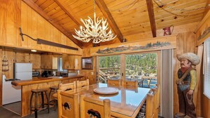 A wooden cabin interior with a dining table, bar stools, a chandelier, and a large window offering a scenic view. - Walking Distance to Boulder Bay! Lakeviews! PET FRIENDLY! Internet! Darts & Poker Table! (Big Bear Lake)