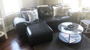 A living room with a black leather sofa, a glass coffee table, and a small side table. - The Bell House | Modernized Bungalow Near Main Street & Hwy 290 Wineries (Fredericksburg)