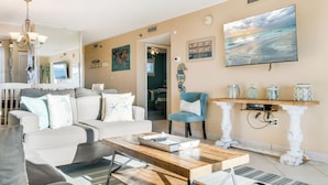 A living room with a sofa, coffee table, and decorative items. There is a television mounted on the wall and a window with blinds. - The Islander 715 | Stunning Panoramic Gulf & Harbor Views | Newly Updated (Destin)