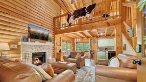 A log cabin interior with a stone fireplace, leather sofas, a dining area, and a kitchen. - ER43 Tulip Tree – 3BR Cabin w/ Hot Tub, Arcade, Pool, & Prime Pigeon Forge Spot (Pigeon Forge)