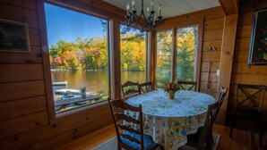 Eine Holzhütte mit Esstisch und Stühlen, große Fenster mit Blick auf einen See und Herbstlaub.