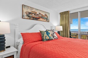 A hotel room with a large bed, two bedside lamps, a painting of boats, a balcony with a view of the sea, and a window with curtains.