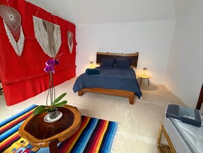 A bedroom with a wooden bed, two bedside lamps, a red wall with macrame decorations, a small round wooden table with a plant, and a colorful striped rug. - Magical jungle Eco Cabana in Tulum Mexico (Macario Gómez)