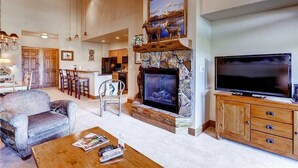 Amplia sala de estar con chimenea de piedra, un televisor de pantalla plana grande y comedor con armarios y sillas de madera.