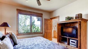 Un dormitorio con una cama grande, un televisor sobre un soporte de madera, una ventana con vista a la vegetación y un ventilador de techo.