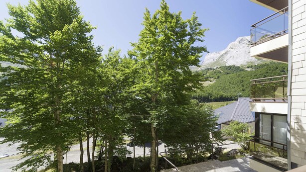 A modern building with a balcony overlooking a mountainous landscape and a parking area with trees.