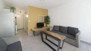 A modern living room with a sofa, a wooden coffee table, a TV, and a kitchen area with a refrigerator and cabinets.