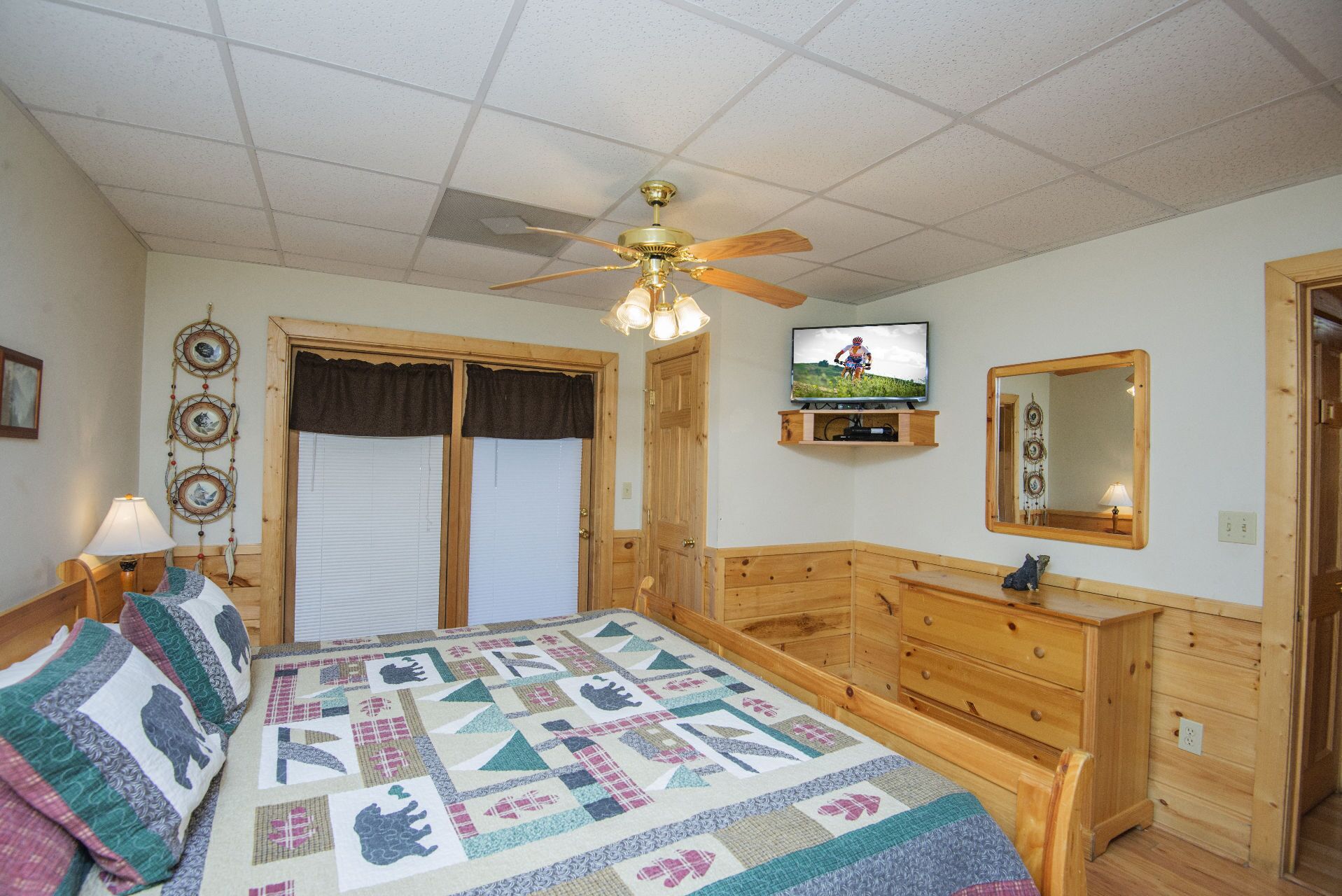 Une chambre avec un lit, une commode en bois, un ventilateur de plafond, une télévision et un attrape-rêves accroché au mur.